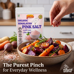 A stand-up pouch of Dinesh Your Mill Organic Himalayan Pink Salt is shown beside a ceramic bowl of roasted vegetables, with a hand sprinkling pink salt crystals over the food. The scene is set on a wooden table with carrots, beets, and other vegetables in a softly blurred kitchen background.